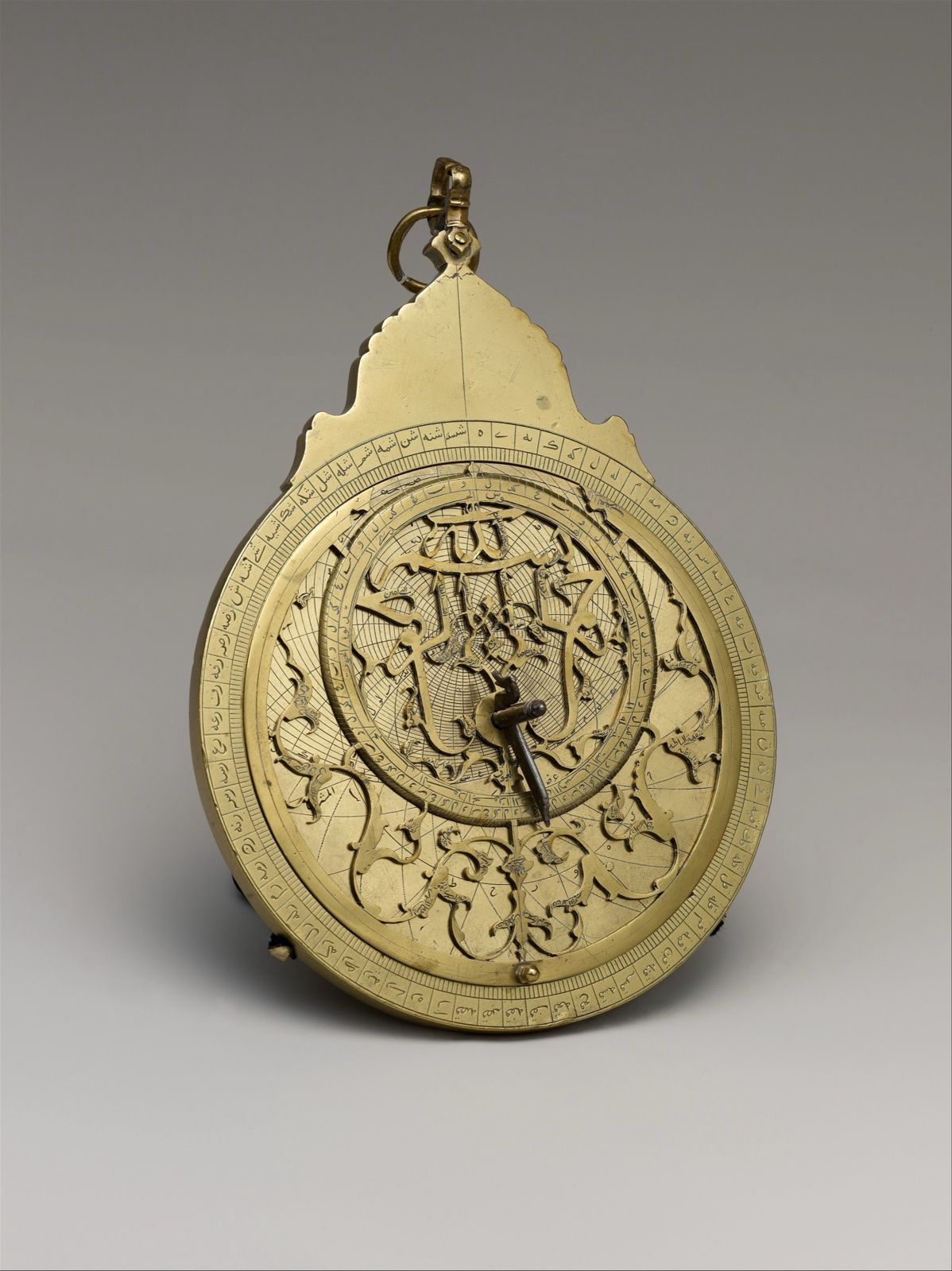 A brass and steel astrolabe, pierced and engraved, hanging from its ring, on a grey ground.