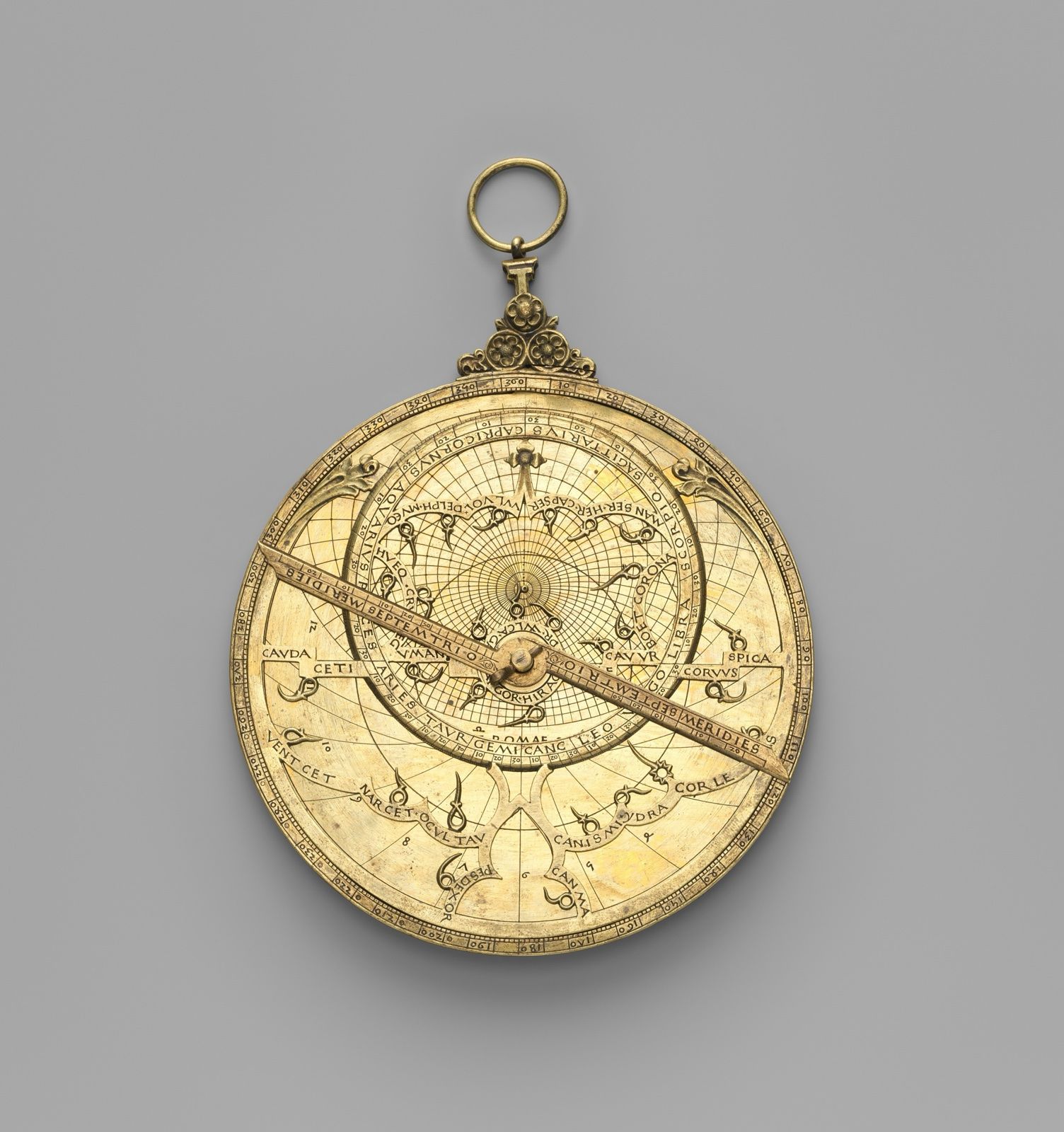 A copper-alloy astrolabe hanging from its ring, an engraved pointer plate turning over a disc of scales, on a grey ground.