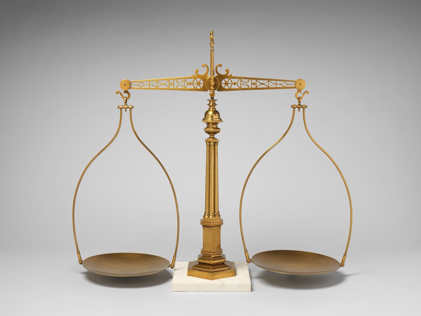 A brass balance on a fluted column and marble base, two pans hanging level from an ornate beam, on a grey ground.