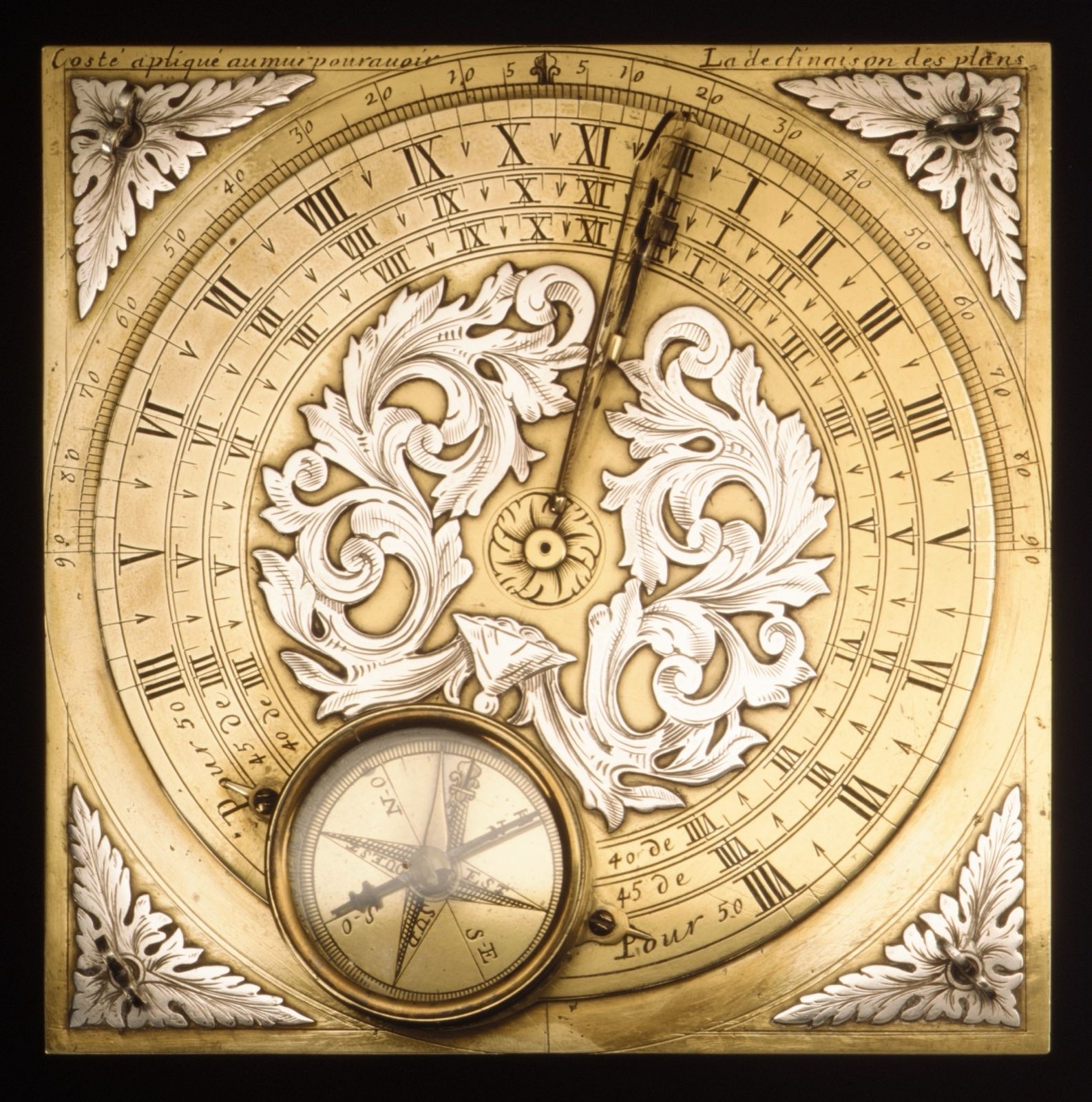 A gilded square plate seen from above: hour scales round its edge, a silver scroll of leaves in the middle and a compass set below.