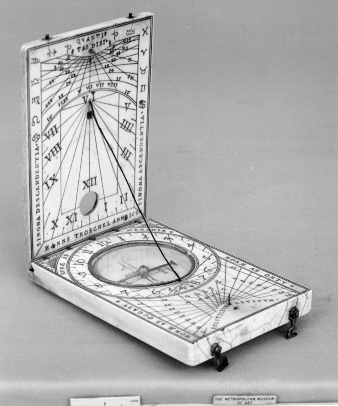 An open ivory and brass folding sundial, its two leaves engraved with hour lines, a small compass set in the lower leaf, on a grey ground.