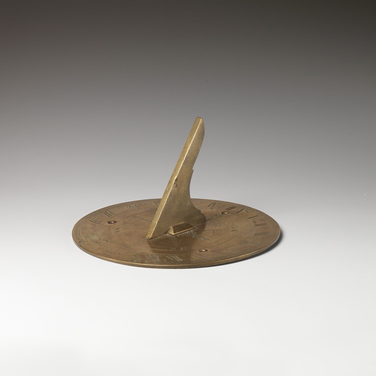 A round brass sundial plate with a tall triangular gnomon casting its edge over the hour lines, on a grey ground.