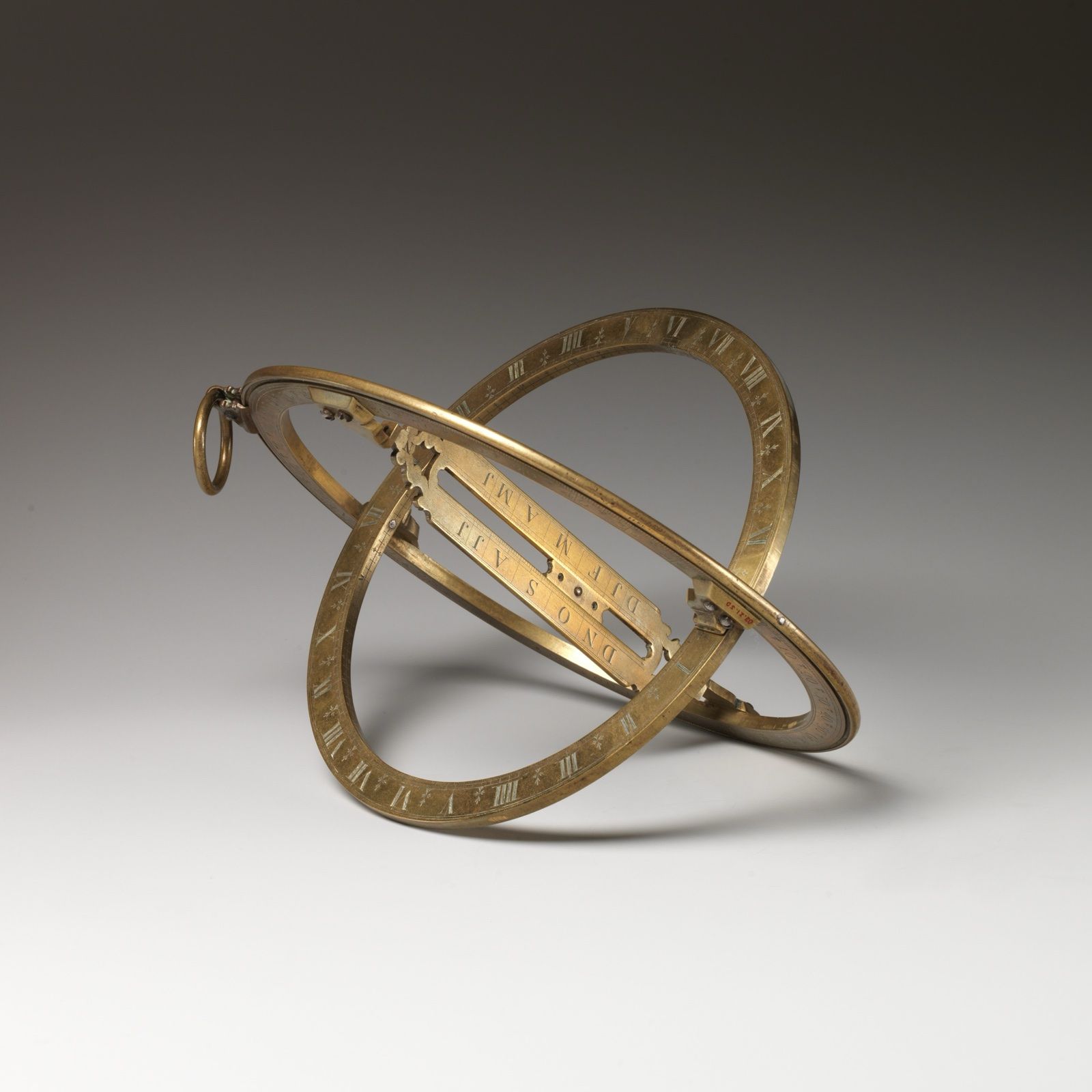Two crossed brass rings, engraved with scales and joined by a sliding bridge, hanging from a small ring, on a grey ground.