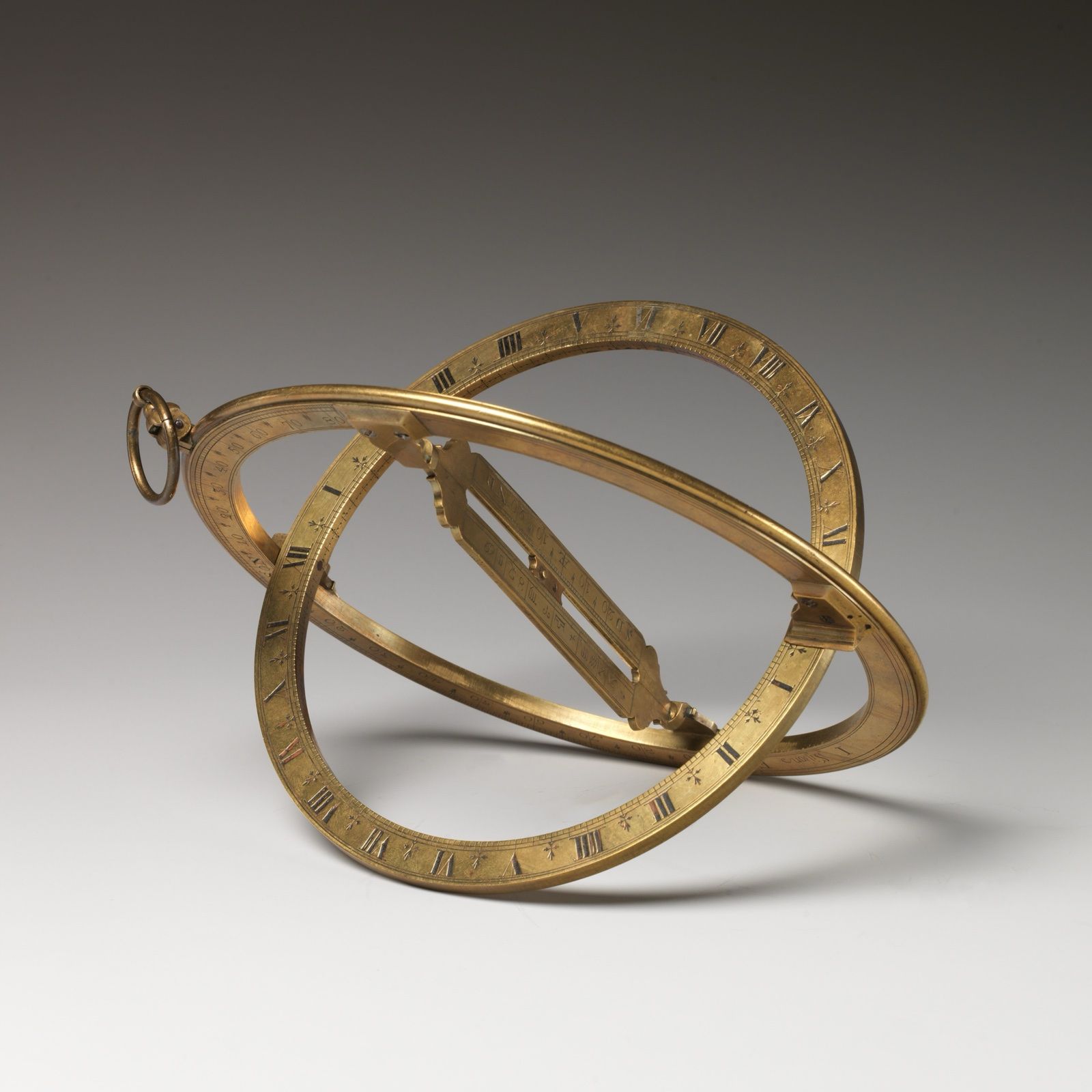 A brass ring sundial opened out, its two engraved rings crossing at right angles around a sliding bridge, on a grey ground.