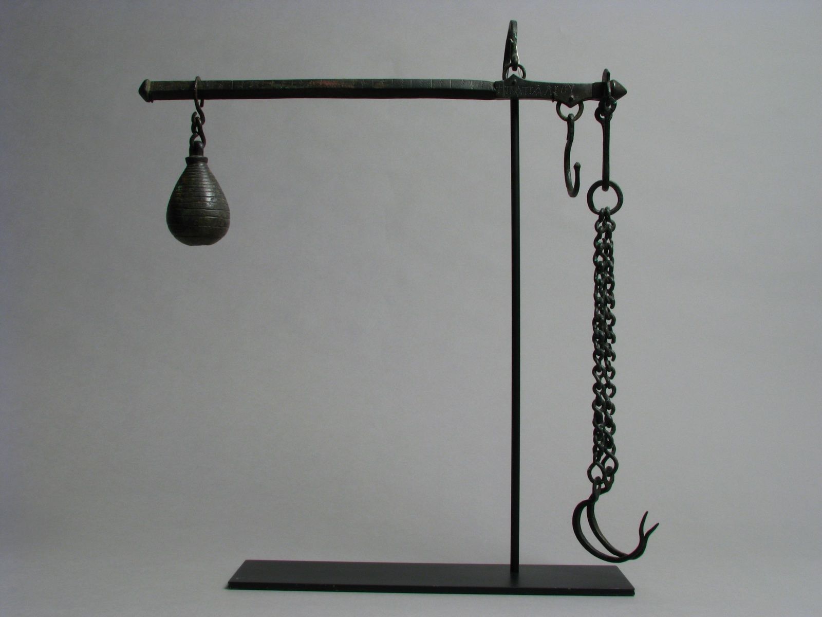 A copper-alloy steelyard hung from a stand: a pear-shaped weight on the long arm, a chain and two hooks on the short one, on a grey ground.