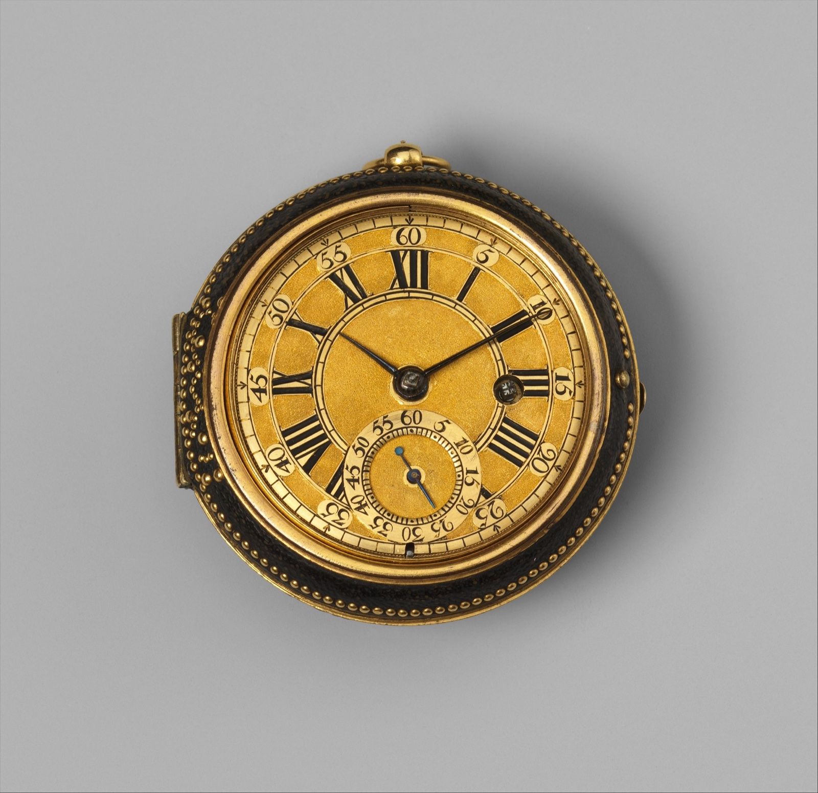 A gold watch dial with black Roman numerals and blued-steel hands, in a dark outer case studded with gold, on a grey ground.